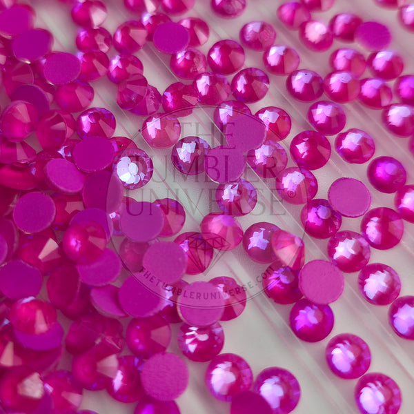 A close-up of many small round pink gemstones or rhinestones scattered on a light surface, with a faint 'The Tumbler Universe' watermark across the center.