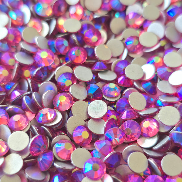 A close-up pile of round, faceted rhinestones with an iridescent pink, purple, and blue finish, showing their silver-toned metal backs. One rhinestone in the center has a visible brand stamp reading the tumbler universe