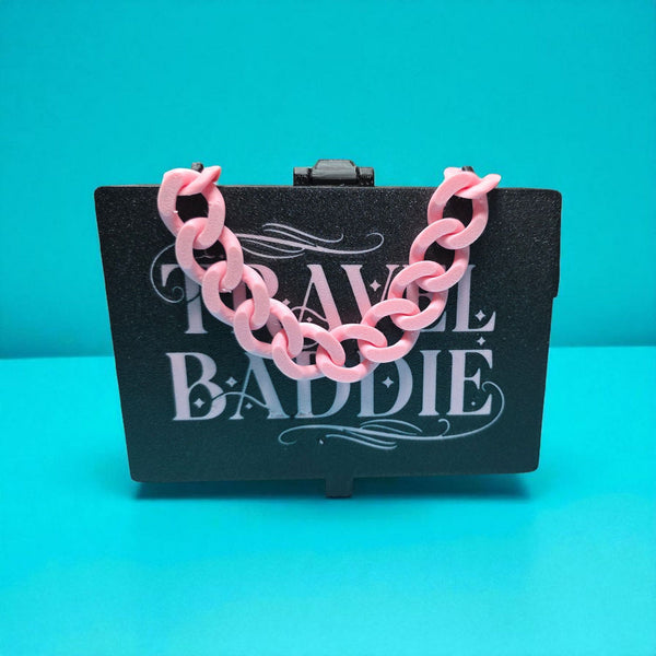 Pink chain necklace on a black card with 'Travel Baddie' text against a teal background