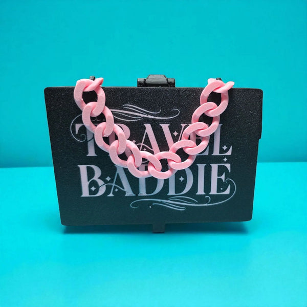 Pink chain necklace on a black card with 'Travel Baddie' text against a teal background