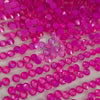 A close-up of many small round faceted gemstones in bright hot pink and magenta tones, arranged on a white surface with a translucent watermark reading 'The Tumbler Universe' across the center.