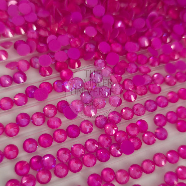 A close-up of many small round faceted gemstones in bright hot pink and magenta tones, arranged on a white surface with a translucent watermark reading 'The Tumbler Universe' across the center.