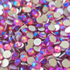 A close-up pile of round, faceted rhinestones with an iridescent pink, purple, and blue finish, showing their silver-toned metal backs. One rhinestone in the center has a visible brand stamp reading the tumbler universe
