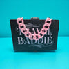 Pink chain necklace on a black card with 'Travel Baddie' text against a teal background
